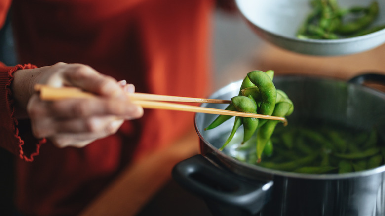A person uses chopsticks to hold cooked edamame over a pot of water