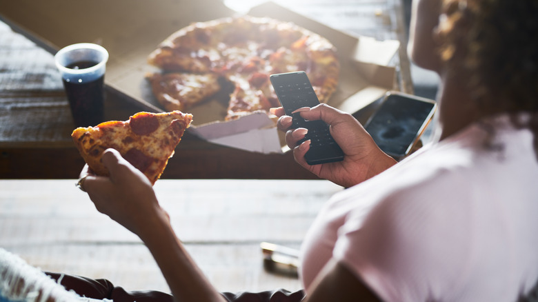 Over-the-shoulder view of a person eating pizza and holding a TV remote