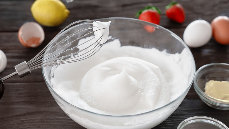 Glass bowl of whipped egg whites with balloon whisk surrounded by eggs, strawberries, and lemon