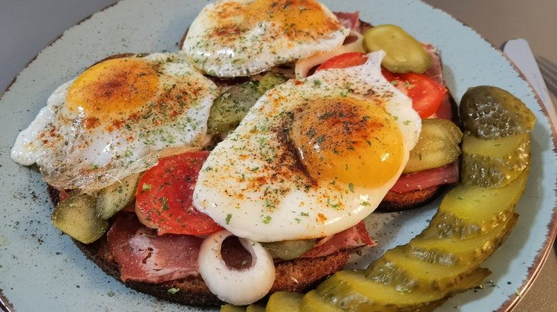 A plated Strammer Max open-faced sandwich with fried eggs, meat, onion, tomatoes, and sliced pickle