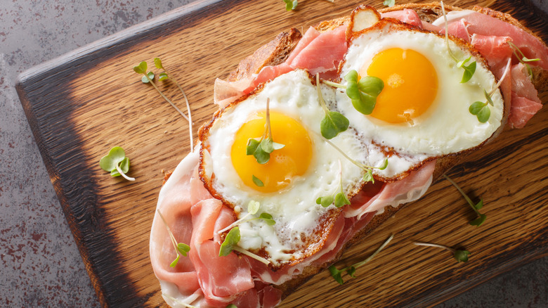 A Strammer Max open-faced sandwich with ham and fried eggs, resting on a cutting board sprinkled with fresh herbs