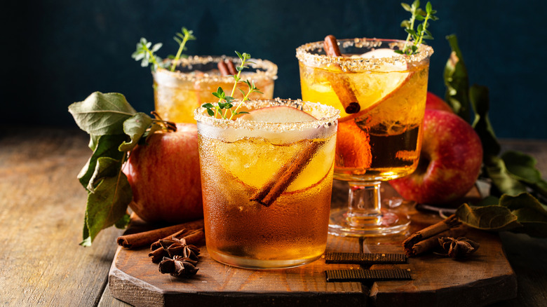 A trio of golden-hued apple cocktails with cinnamon stick and apple slice garnishes and displayed with whole apples, star anise, and cinnamon sticks on a wooden table