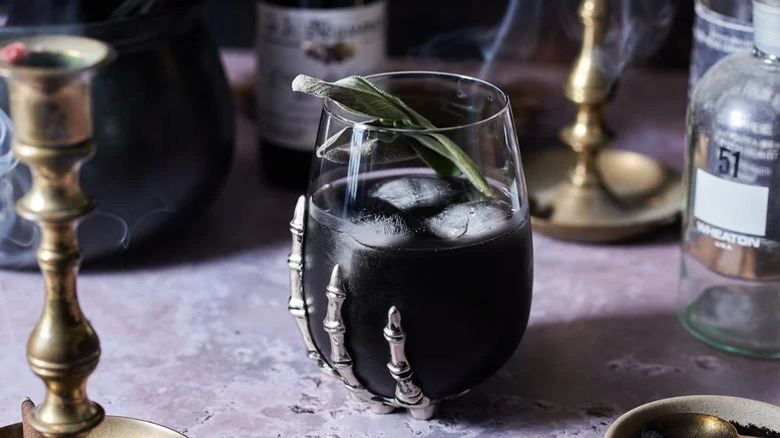 A black drink with ice and a sprig of sage in a glass with metallic skeleton fingers reaching up from the bottom.
