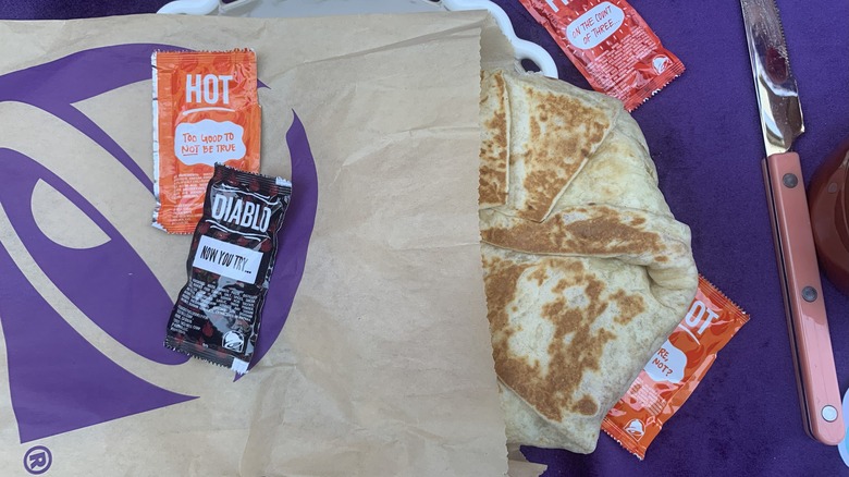 Brock's California Crunchwrap half covered by a to-go bag and surrounded by Taco Bell sauce packets