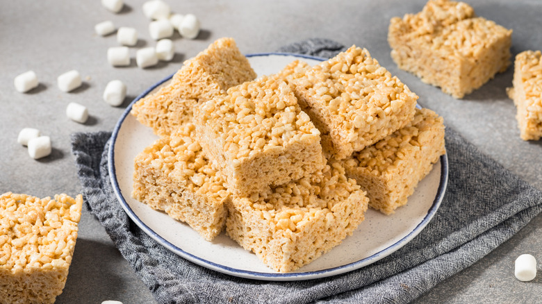 A pile of Rice Krispies treats on a white plate, with several Rice Krispies treats and marshmallows scattered around it