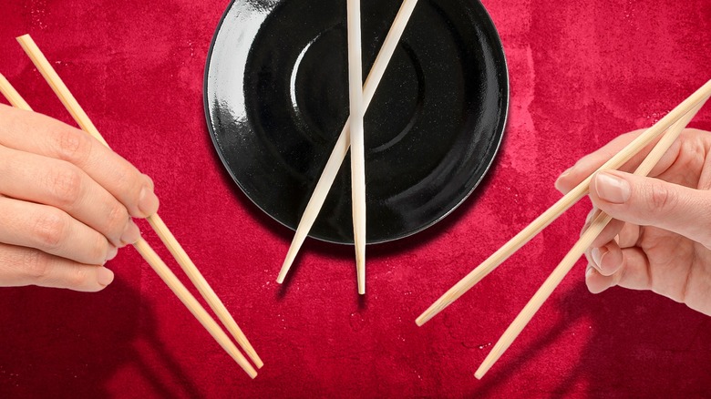two hands holding chopsticks and one pair crossed on a plate