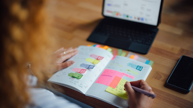 Person checking their daily planner with sticky notes in front of a laptop