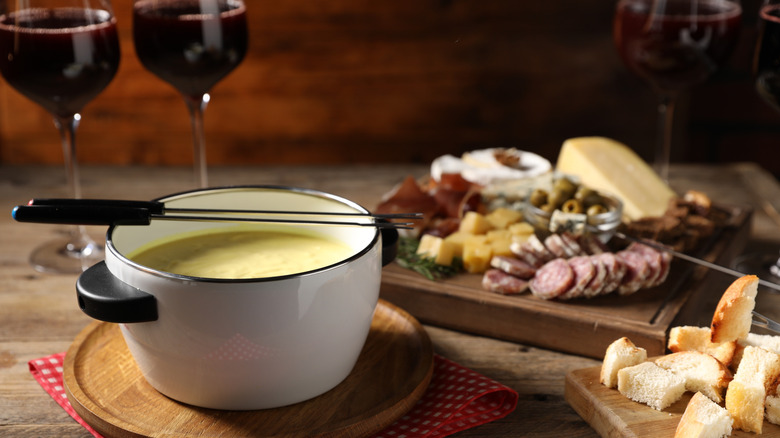 fondue pot on table filled with melted cheese plus a charcuterie board, bread cubes, and glasses of red wine besides
