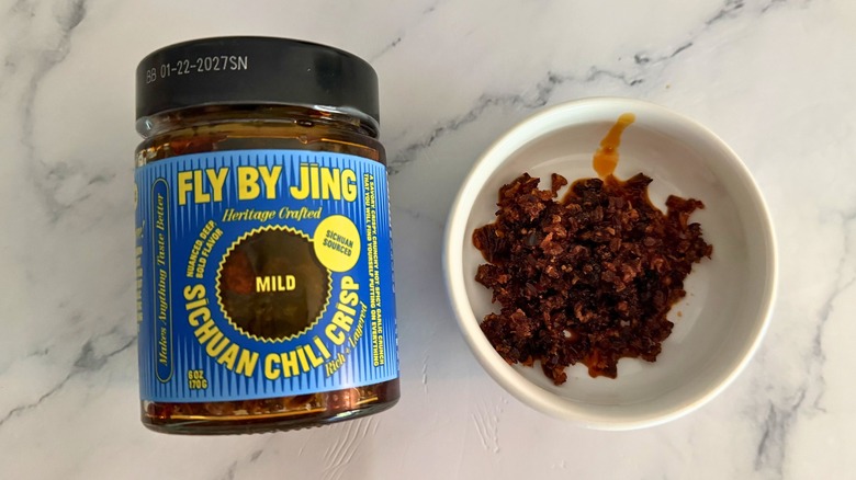 Container of Fly By Jing Mild Chili Crisp next to small white bowl of chili crisp on marble surface