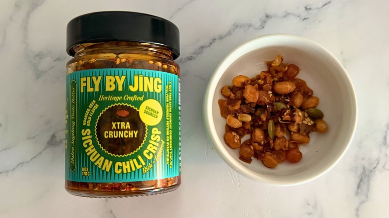 Container of Fly By Jing Xtra Crunchy Chili Crisp next to small white bowl of chili crisp on marble surface