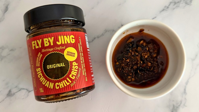 Container of Fly By Jing Original Chili Crisp next to small white bowl of chili crisp on marble surface