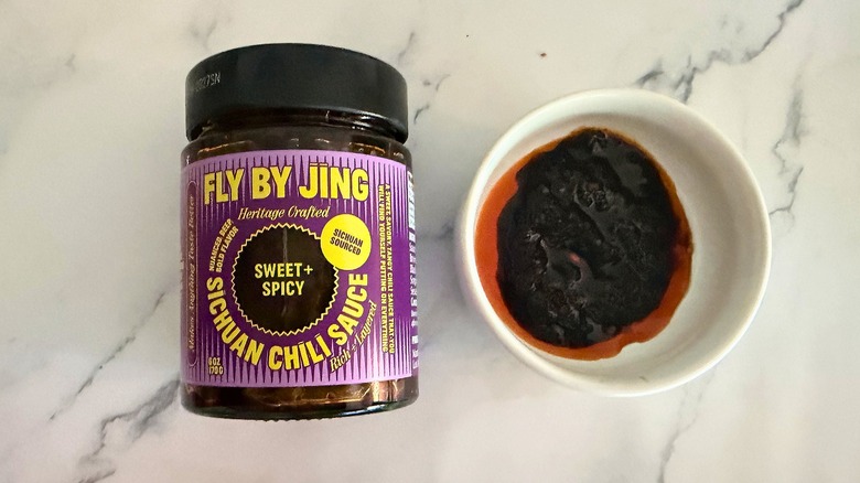 Container of Fly By Jing Sweet & Spicy Chili Crisp next to small white bowl of chili crisp on marble surface