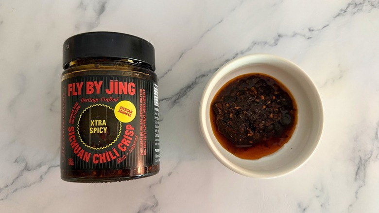Container of Fly By Jing Xtra Spicy Chili Crisp next to small white bowl of chili crisp on marble surface