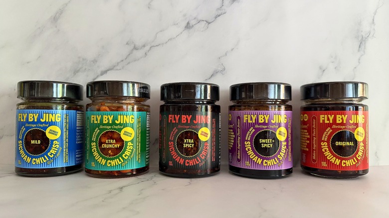 Five containers of different Fly By Jing chili crisps on marble background