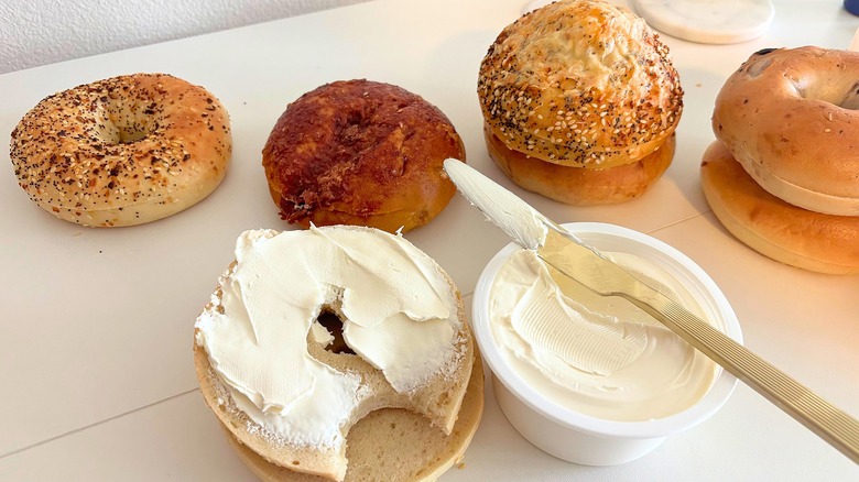 Bagels on table behind bagel with cream cheese, tub of cream cheese, knife with cream cheese