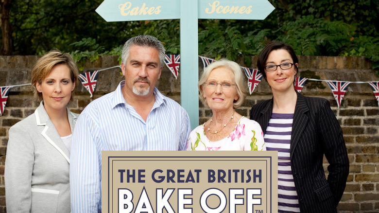 Four hosts from The Great British Bake Off stand together in front of a signpost labeled "Cakes" and "Scones," smiling for the camera.