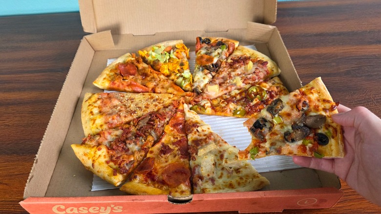 A slice of each pizza flavor in a pizza box with a hand holding a veggie pizza slice