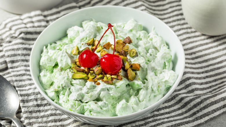 A white bowl on a striped tablecloth full of fluffy, pale green Watergate salad topped with crushed pistachios and two maraschino cherries