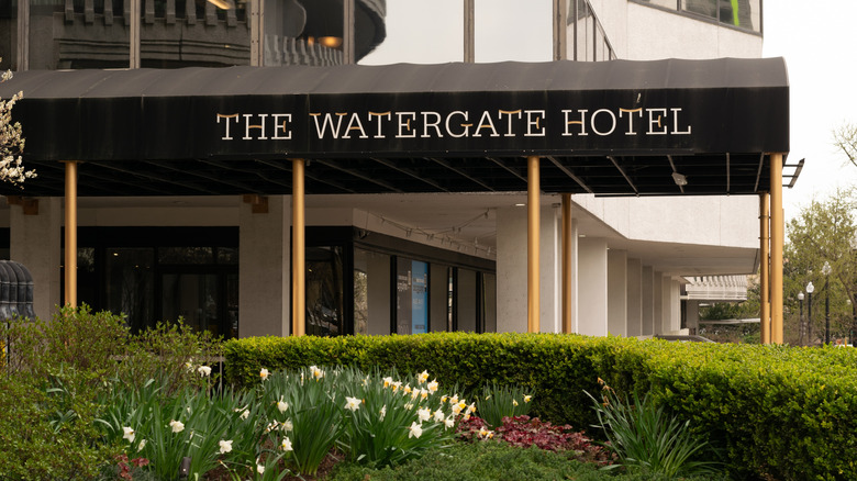 A canopy outside the Watergate Hotel featuring the name, located behind a small, landscape garden with bushes and flowers