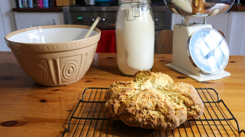 a craggy loaf of Irish soda bread, on a cooling rack on a wooden counter, with a mixing bowl and spoon, a jar of baking soda, and a baker's scale shown behind it