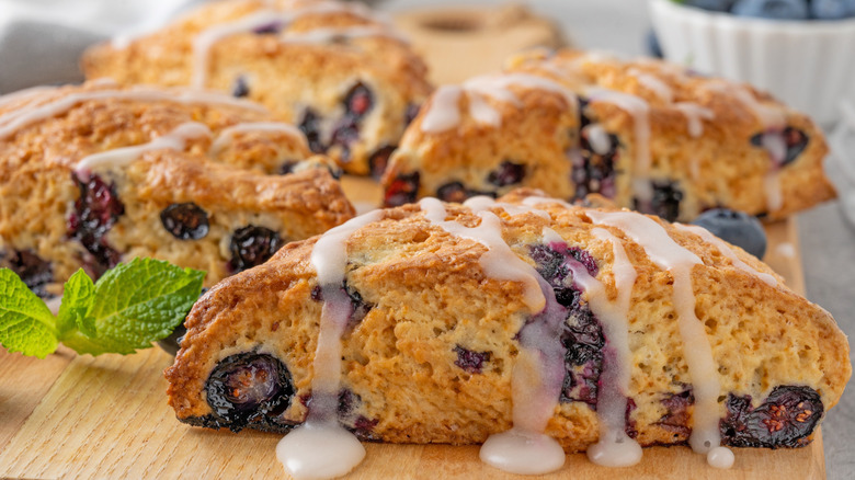 blueberry scones with lemon glaze, shown on a wooden cutting board, garnished with fresh blueberries and mint leaves, with a white ramekin of additional berries and mint visible but out of focus to the rear