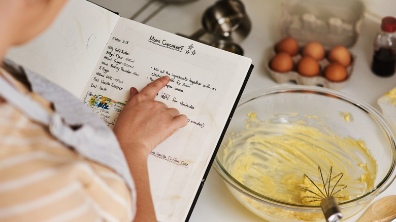 woman consulting a hand-written recipe titled "Mom's Cupcakes?," with a mixing bowl, ingredients and measuring cups on the counter in front of her