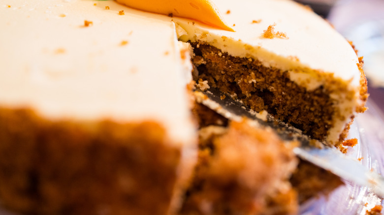 closeup image of a rich and crumbly carrot cake with cream cheese icing, with one large wedge cut out and the slicing knife remaining in the empty spot left by the wedge of cake
