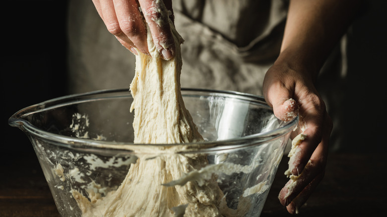 a woman's hands pulling a strand of soft and sticky bread dough from a glass mixing bowl, as if evaluating its texture