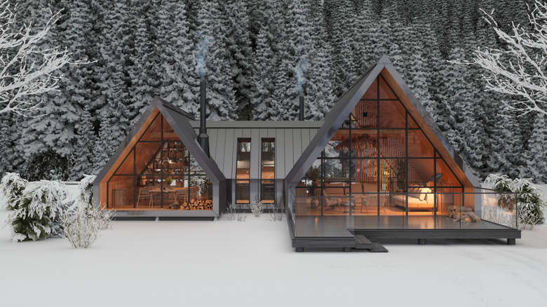 Image of a very modern mountainside chalet, of two A-frame structures connected at the rear, surrounded by snowy conifers and with smoke coming from its twin chimneys