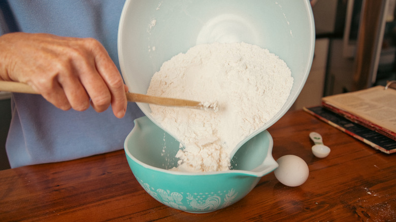 hands pouring flour from one glass mixing bowl to another, with a nearby measuring spoon implying that a secondary ingredient has been added