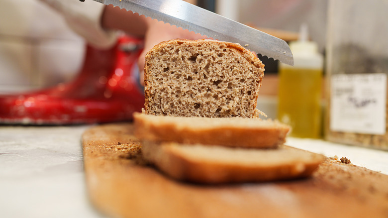 home baker cutting slices from a fresh loaf of whole wheat bread with a serrated knife, with a red tilt-head stand mixer visible but out of focus in the background