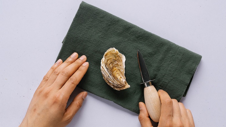 Top-down shot of oyster on green napkin, hand holding knife beside