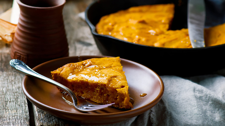 Slice of cornbread with melted butter on brown plate with fork with black skillet of cornbread in background.