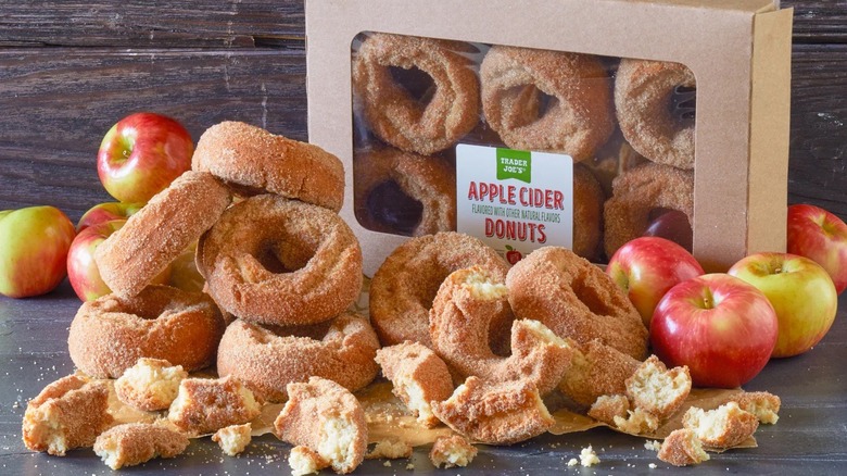 A pile of Trader Joe's Apple Cider Donuts and juicy red apples in front of a box of six donuts