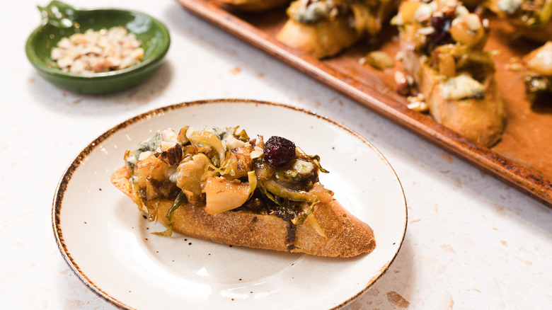 Plate with a slice of bruschetta with Brussels sprouts, pears and gorgonzola cheese, plus more on a baking sheet in background