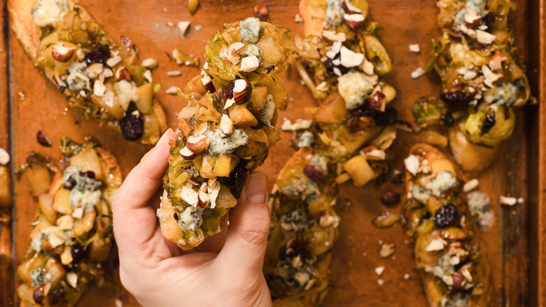 Hand holdinga slice of bruschetta with Brussels sprouts, pears and gorgonzola cheese, over a a baking sheet