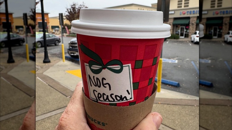A person holding a red Starbucks holiday cup that says "Nog Season" on it