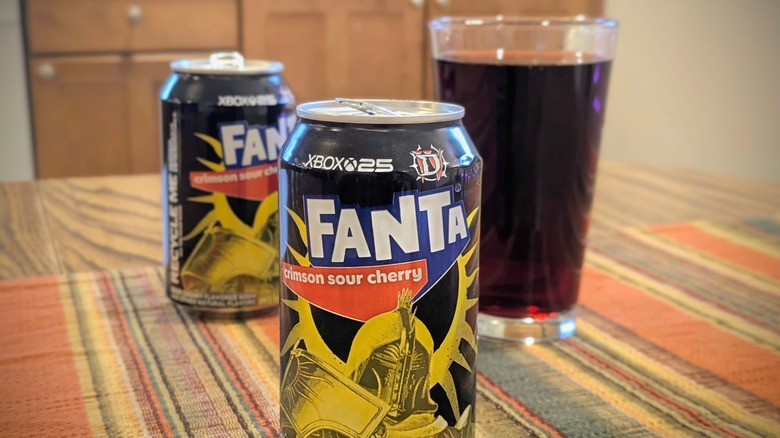 A can of Fanta Crimson Sour Cherry with another can and a glass of soda in the background