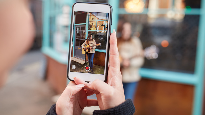 Hands holding a phone that's filming a video of someone playing guitar in front of a building