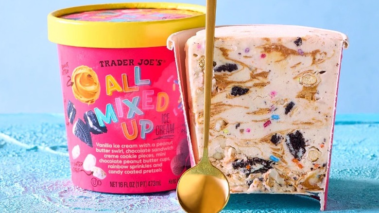 Two pints of Trader Joe's All Mixed Up Ice Cream with one cut in half and having a golden spoon in front, showing the inside, and a whole closed pint on a teal background