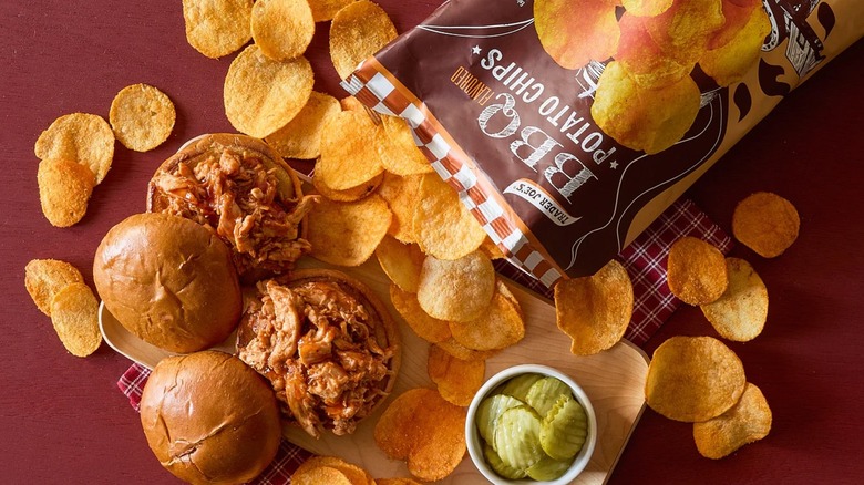 An open bag of Trader Joe's BBQ Flavored Potato Chips scattered around a ramekin of pickles and two barbecue sandwiches
