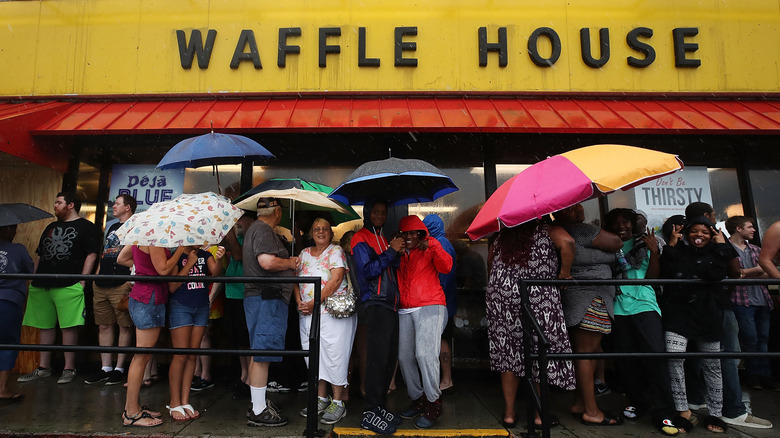 People wait outside of a Waffle House in stormy weather holding umbrellas.