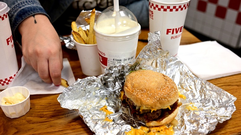 Customer sitting at table in Five Guys restaurant with cheeseburger, milkshake, cup of fries, napkins, and drink cups