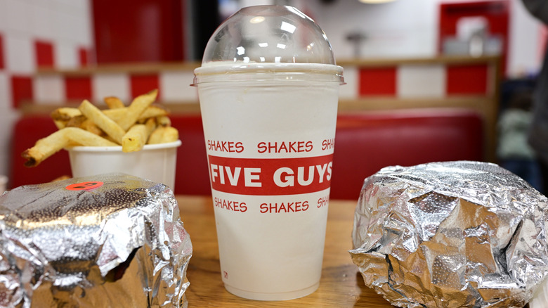Five Guys cup with red logo on table next to cup of fries and two burgers wrapped in foil