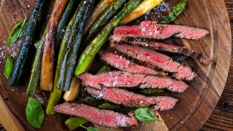 A grilled, sliced flat iron steak sits on a wood plate next to grilled asparagus.