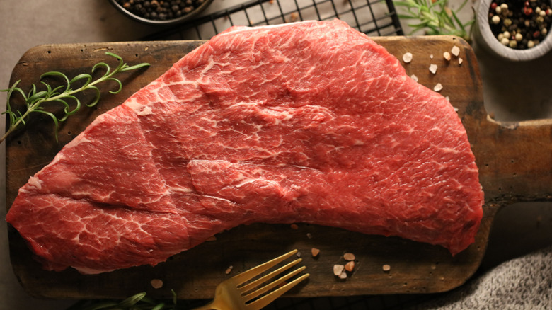 A raw flat iron steaks on a cutting board with salt, peppercorns and a sprig of rosemary