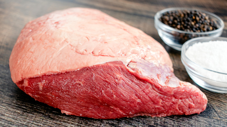 A big, raw rump roast sits on a wooden counter beside salt and pepper seasonings.