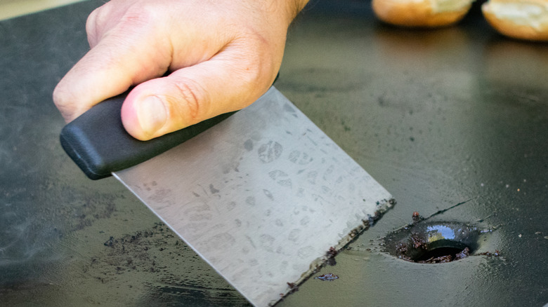 Grease being scraped off of a flat-top grill with a bench scraper