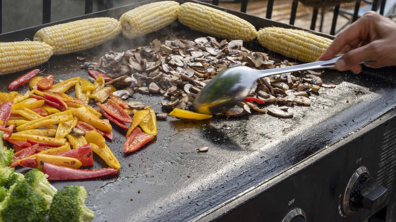 Mushrooms, peppers, corn, and broccoli cooking on an outdoor flat-top grill