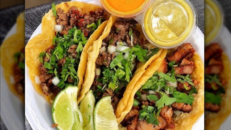 Three tacos with lime wedges and orange and green sauce on the side